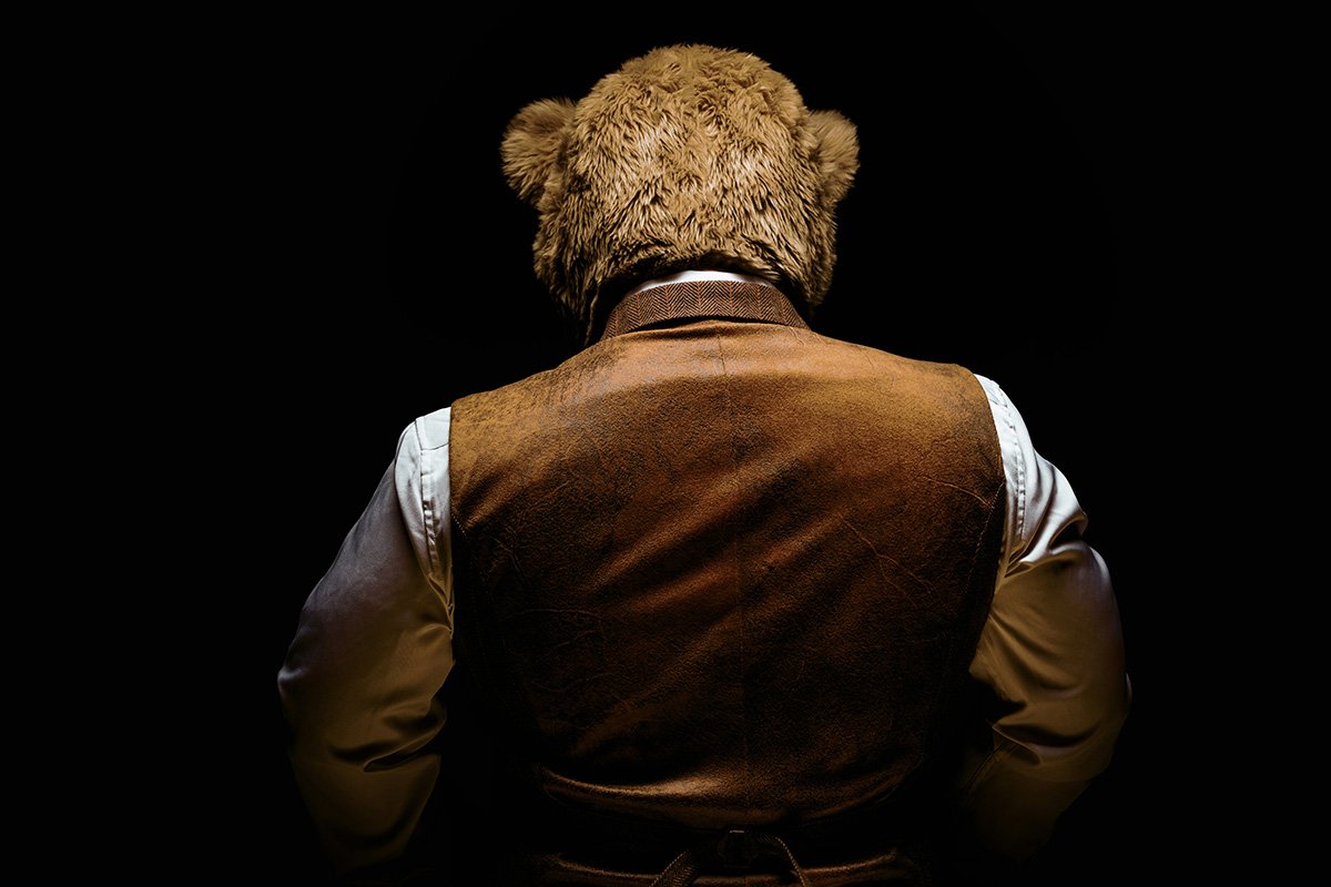 A shadowy photo of a person in a bear costume and leather vest facing away from the camera so thier back is to us.