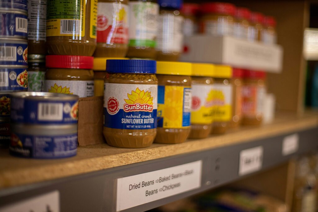 A close-up photo with shallow depth of field showing a shelf in a food pantry with peanut butter and canned fish.