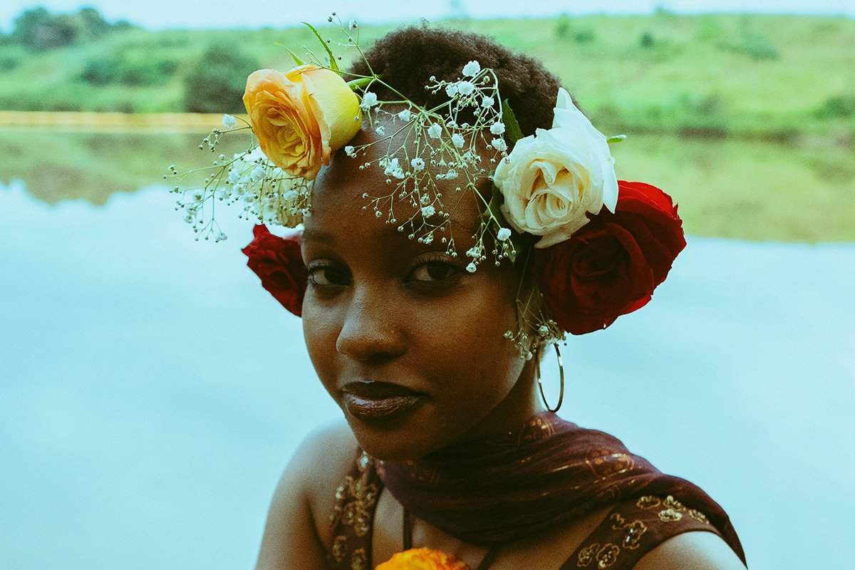 A Black woman with a rose and baby’s breath flower crown sits before a body of water and looks into the camera with a relaxed look on her face.