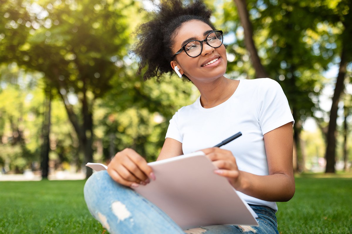 A young Black girl wearing glasses sits on a field of grass with a smile on her face. She writes in a notepad.
