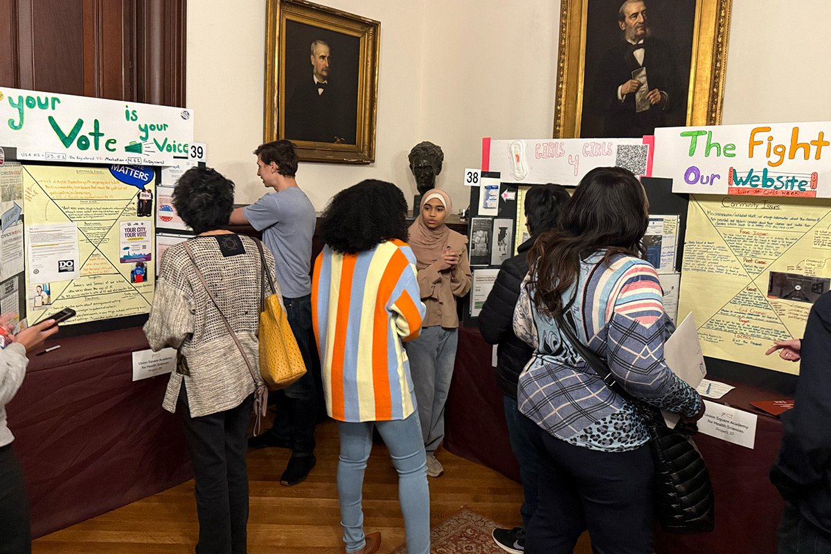 A corner of a wood-finished room is filled with tri-board projects as people read the poster content.