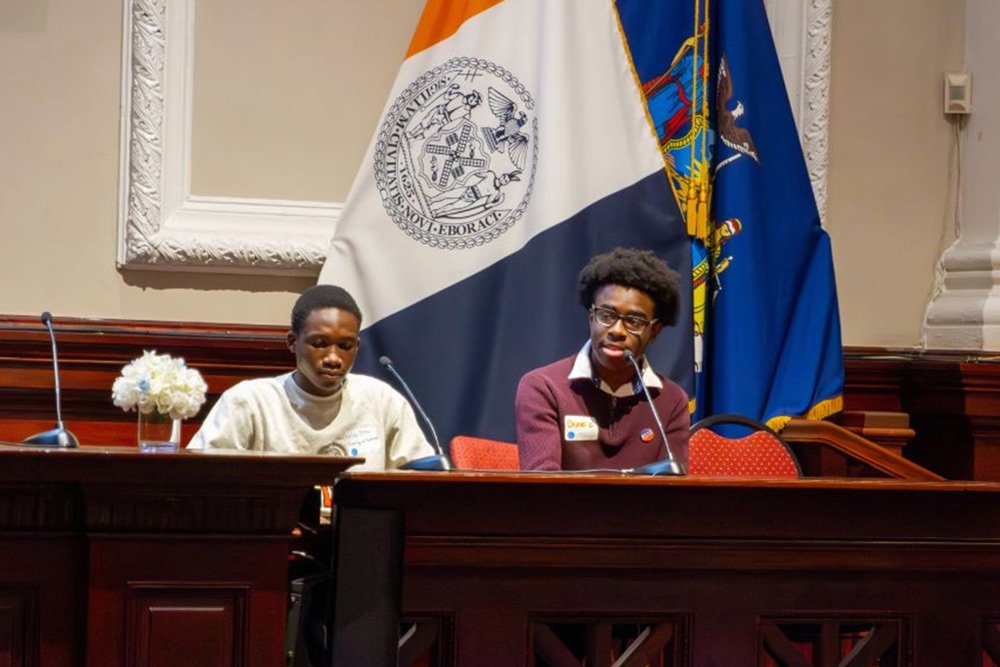 Photo of interviewee, Bismark Ghanney, a Black young man with a textured Afro, glasses, and maroon shirt, sitting behind a microphone in a wooden bench.