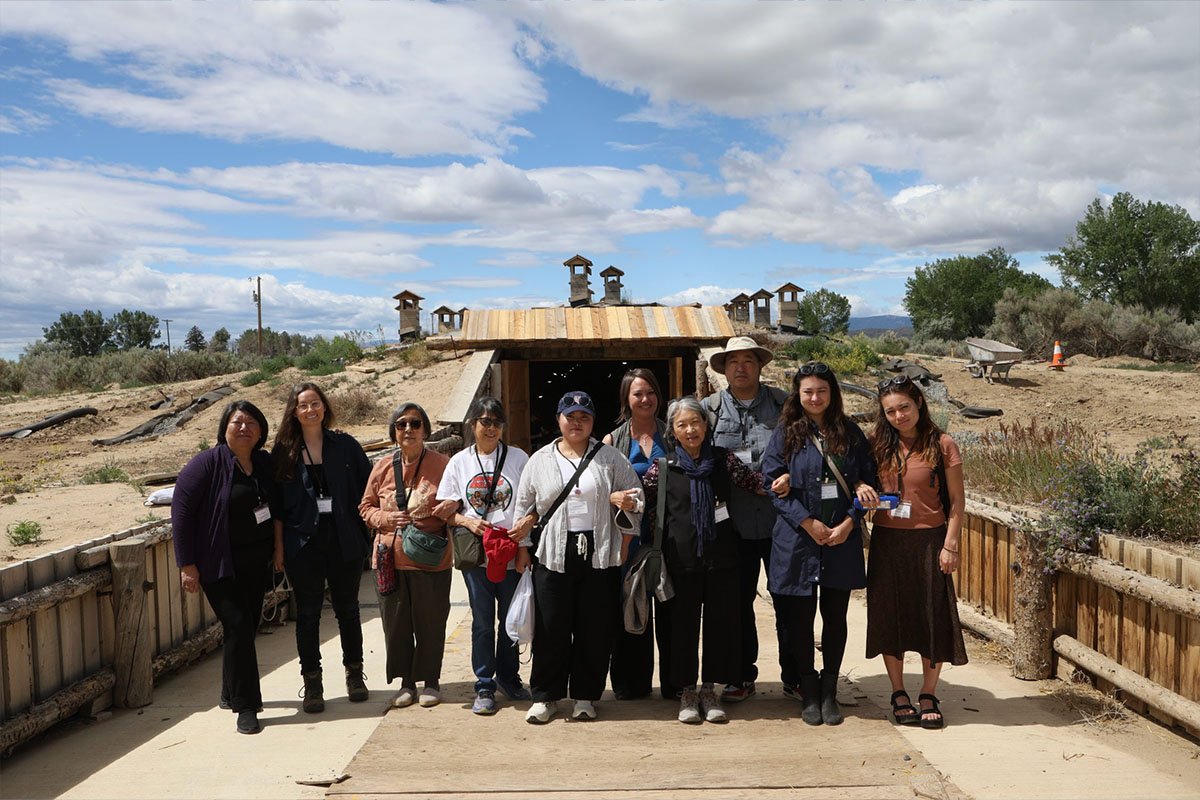 A group of East Asian and East Asian American people of varying ages stands on a paved road in front of a wooden structure. Heart Mountain, Wyoming.