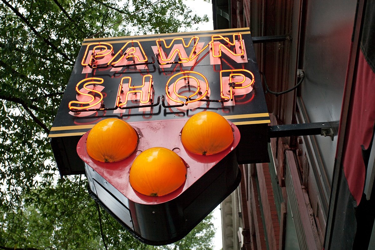 An upward view of a neon sign reading “Pawn Shop” attached to a wooden panel on the side of a building.