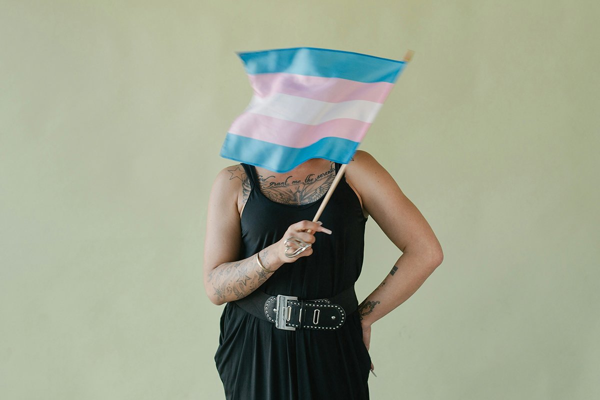 A person stands with one hand on their hip and the other waving a trans flag in front of their face.