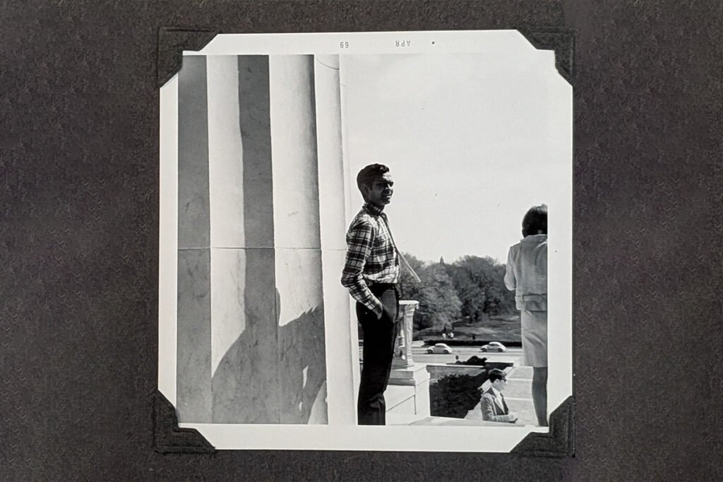 A Black and White photo of a Brown man wearing a palid shirt and slacks, standing at the Lincoln Memorial with his hands in his pocket.