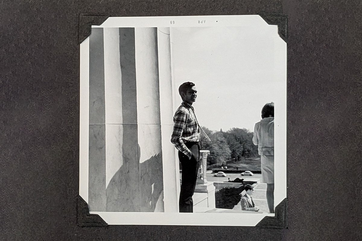 A Black and White photo of a Brown man wearing a palid shirt and slacks, standing at the Lincoln Memorial with his hands in his pocket.