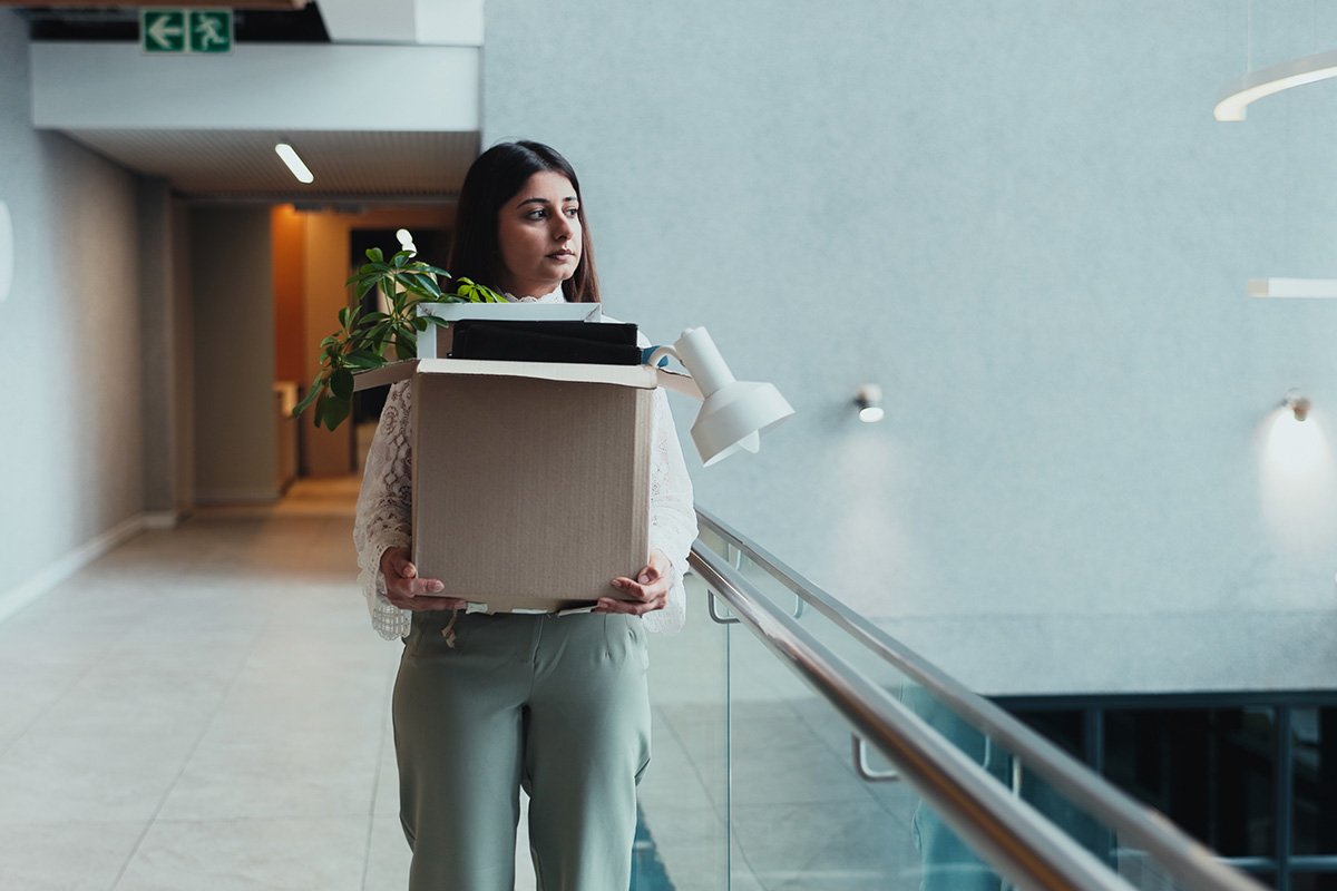 A young, brown woman with dark hair walks down a hallway in an office building. She is holding a box with her belongings, including some documents and a plant. She has been fired.