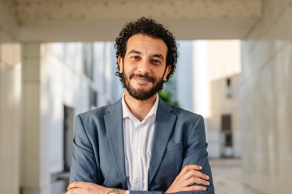 A Middle Eastern man with curly hair and a beard wears a professional-looking suit and stands with his arms crossed in an office hallway.