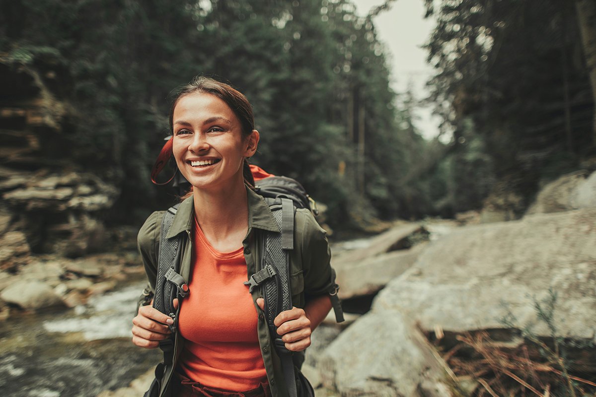 A woman with dark brown hair in a ponytail smiles on the banks of a rocky river in the forest. She has a backpack on her back.