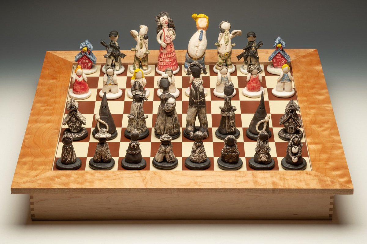 A custom-made chess board where many of the pieces are symbols of white capitalism and violence in America-an armed gunman, KKK members, kneeling children, bishops with money in their pockets, and faceless Native children.