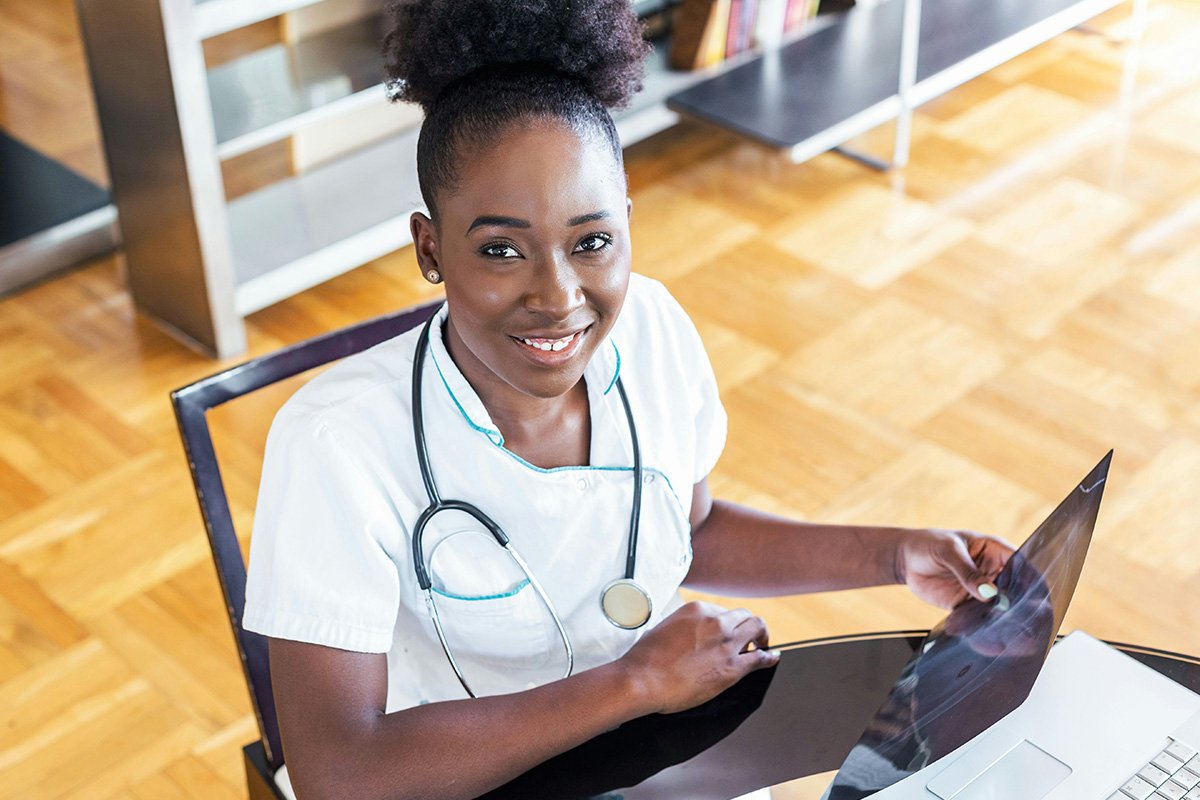 A Black woman in a white lab coat sits in a desk chair while holding an x-ray a smiling up at the camera