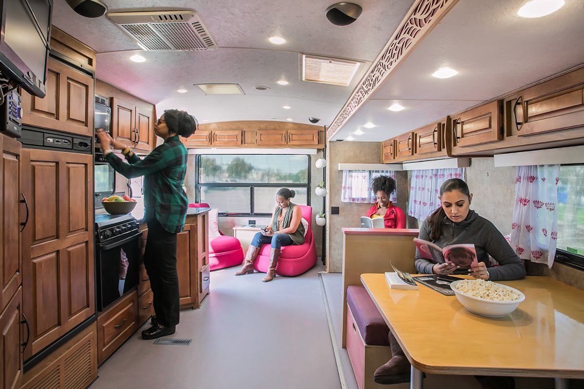The interior of a mobile home which has been refinished with medium-toned wood and pink furniture. Several, diverse women occupy the room sitting, reading, and using the microwave.