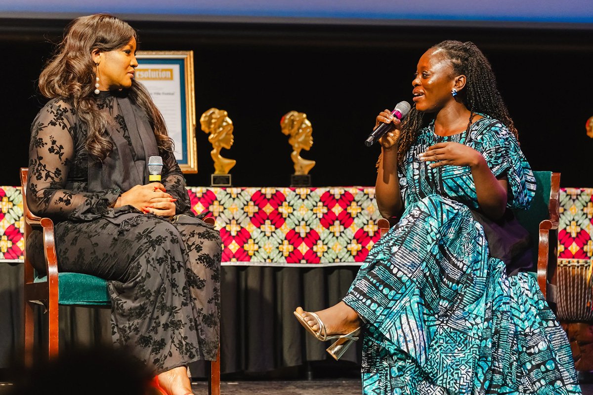 Nigerian Actress Omotola Kehinde sitting on a stage with long black hair, and a black dress. She sits across from a woman wearing a blue and white Kente patterned dress. Behind them is kente cloth-covered table topped with golden awards.