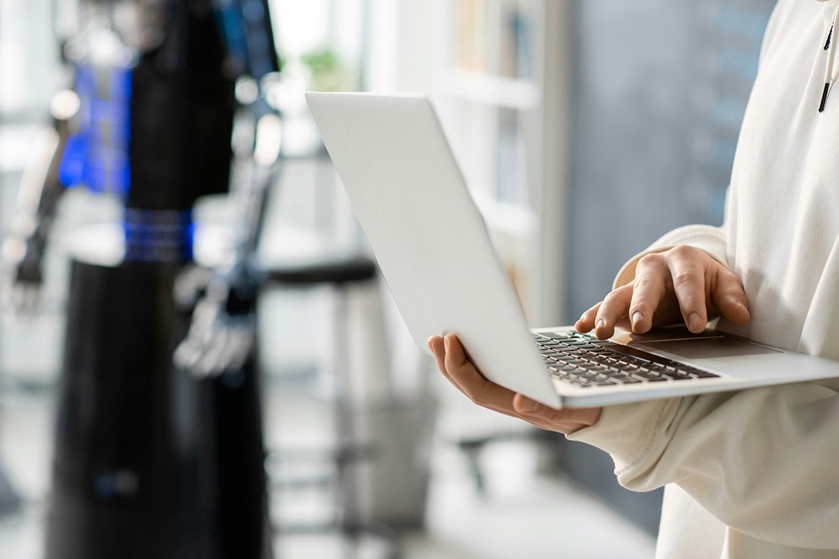 A person’s hands type on a laptop while a standinf robot looms in the background, just out of focus.