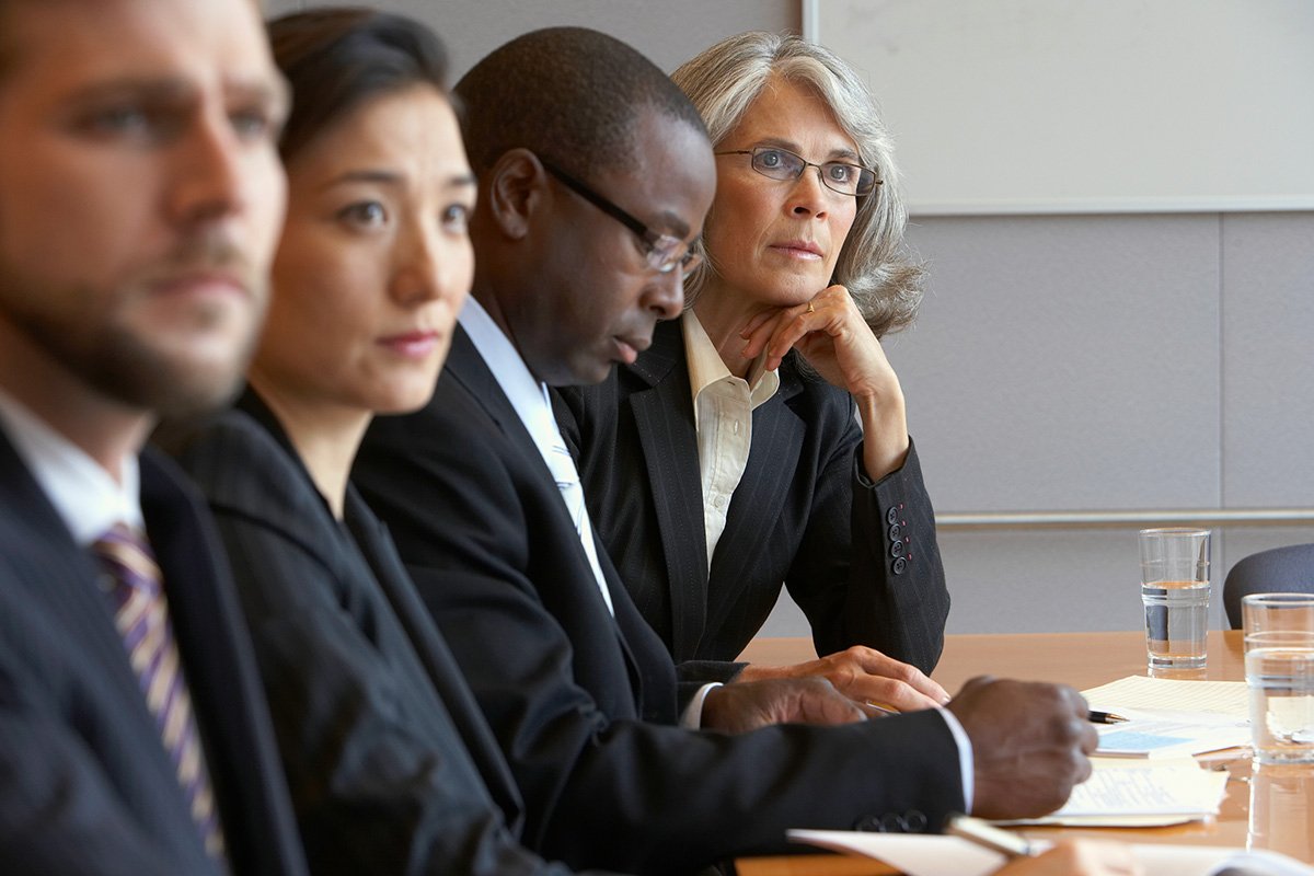 A group of four diverse adults in black suits sitting at an offic emeeting table and looking in the same direction.