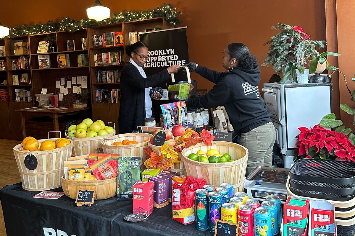 A Black woman stands behind the counter of an indoor farm stand, while handing a shopping basket to a customer. A sign reading “Brooklyn Supported Agriculture” is in the background.