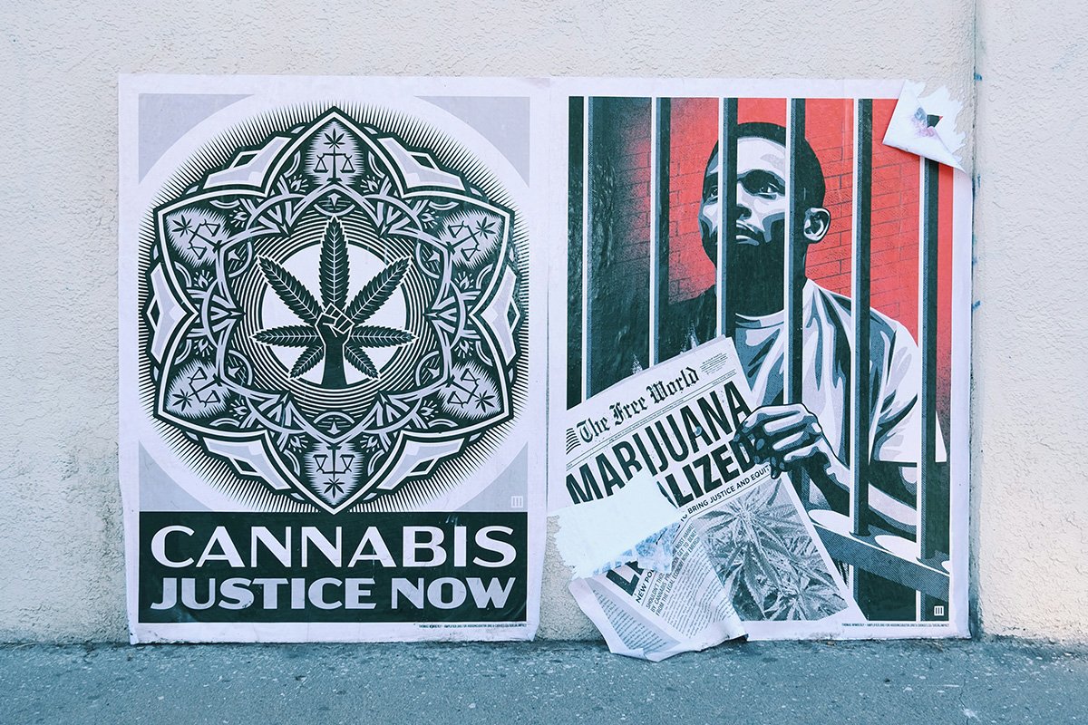A mural on a sidewalk wall, with a Black man behind jail bars holding a newspaper called “The Free World” with a headline reading “Marijuana Legalized”. Beside him are the words “Cannabis Justice Now” with a mandala with images of justice scales, marijuana leaves, and a power fist.