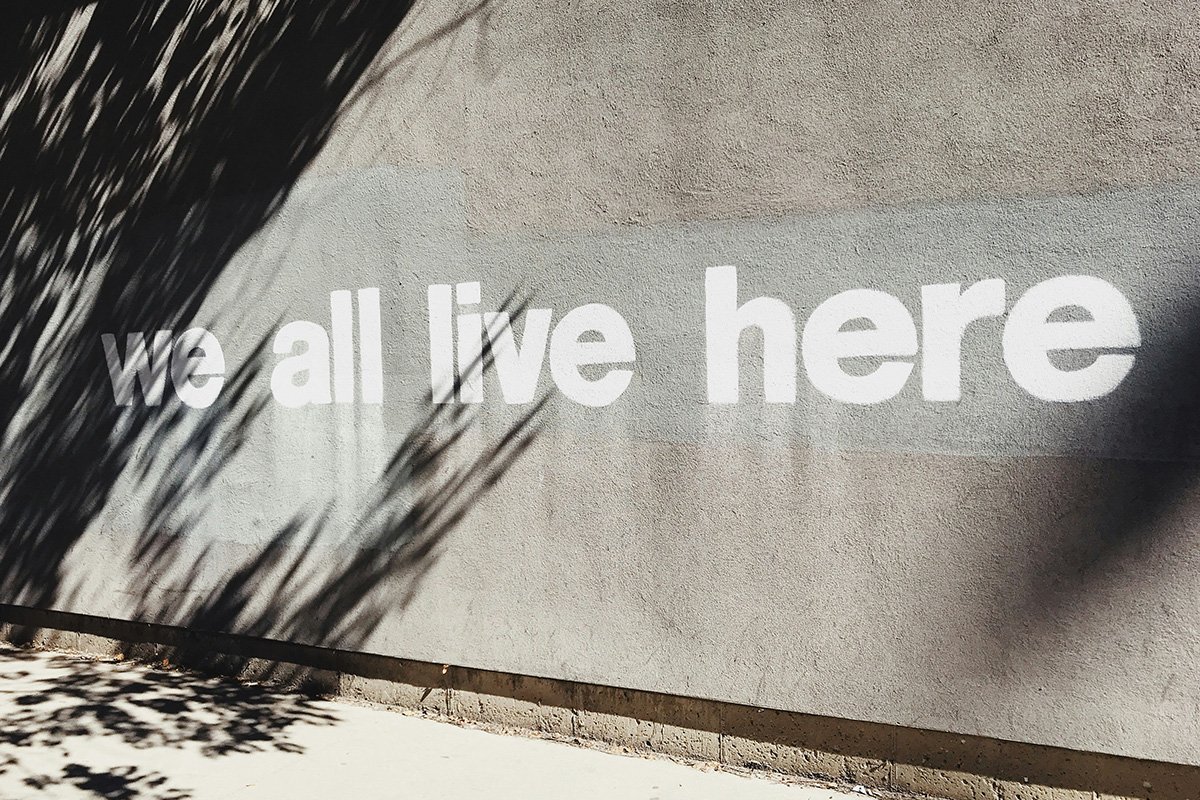 Stenciled words graffiti’d on the side of a buidling reading, “we all live here”