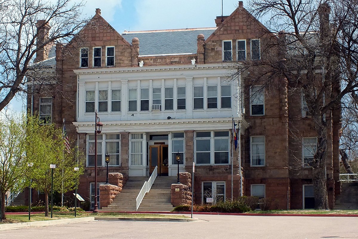 The Colorado School for the Deaf and Blind in Colorado Springs, Colorado. A two story Romanesque Revival building made of large, red stone masonry with two cross gables and a wooden white center with bay windows.