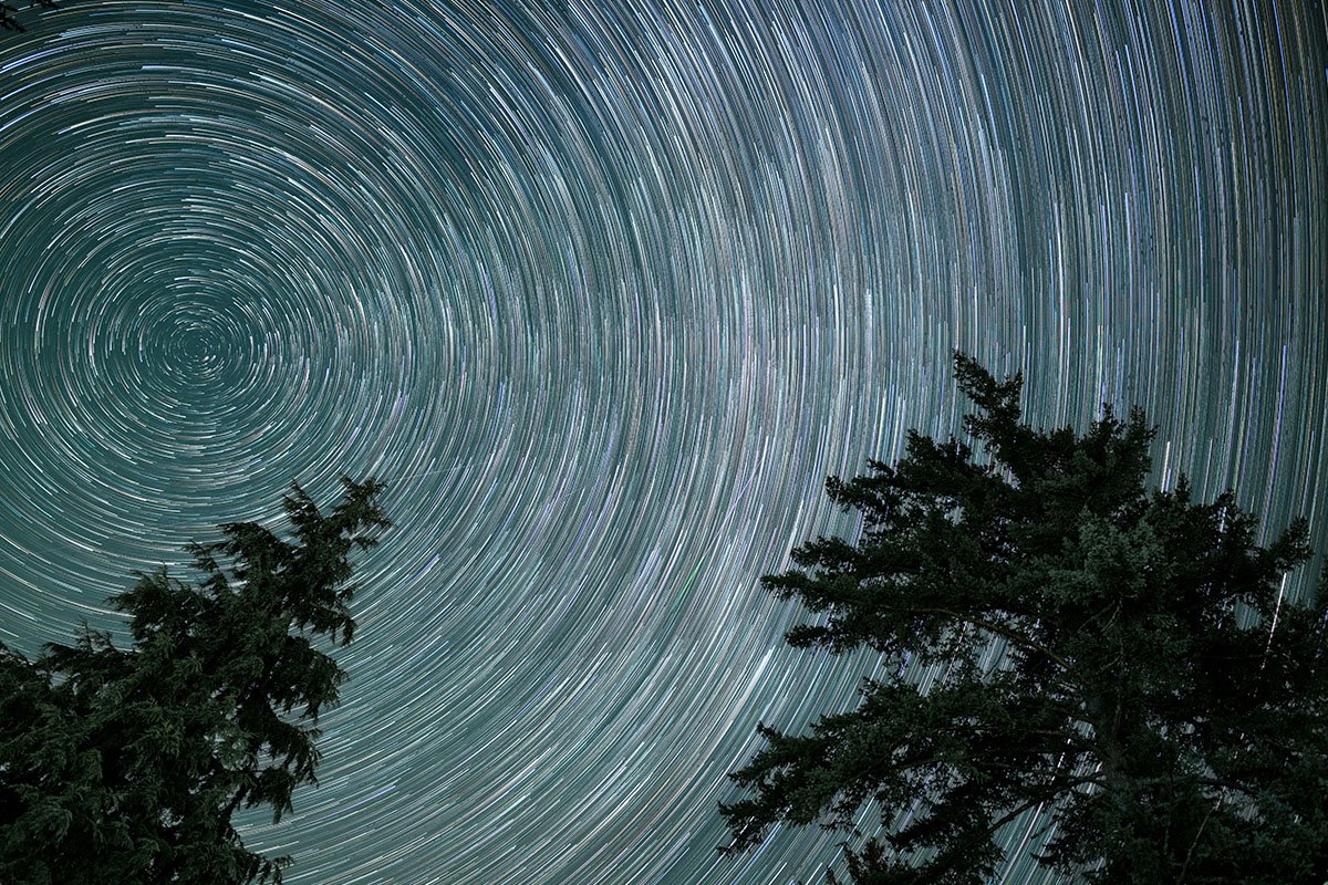 A timelapse of many stars in the sky, creating a spiral affect around a central star in the sky. Trees line the bottom border of the image.