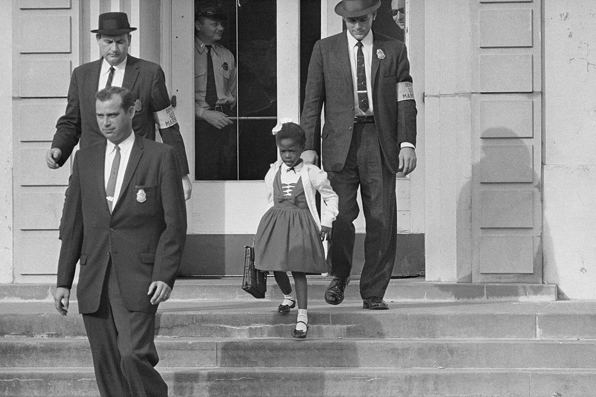 A black and white photo of Ruby Bridges–a young Black girl with a cardigan and hairbow–with three suited US Marshals. William Frantz Elementary School, New Orleans, 1960