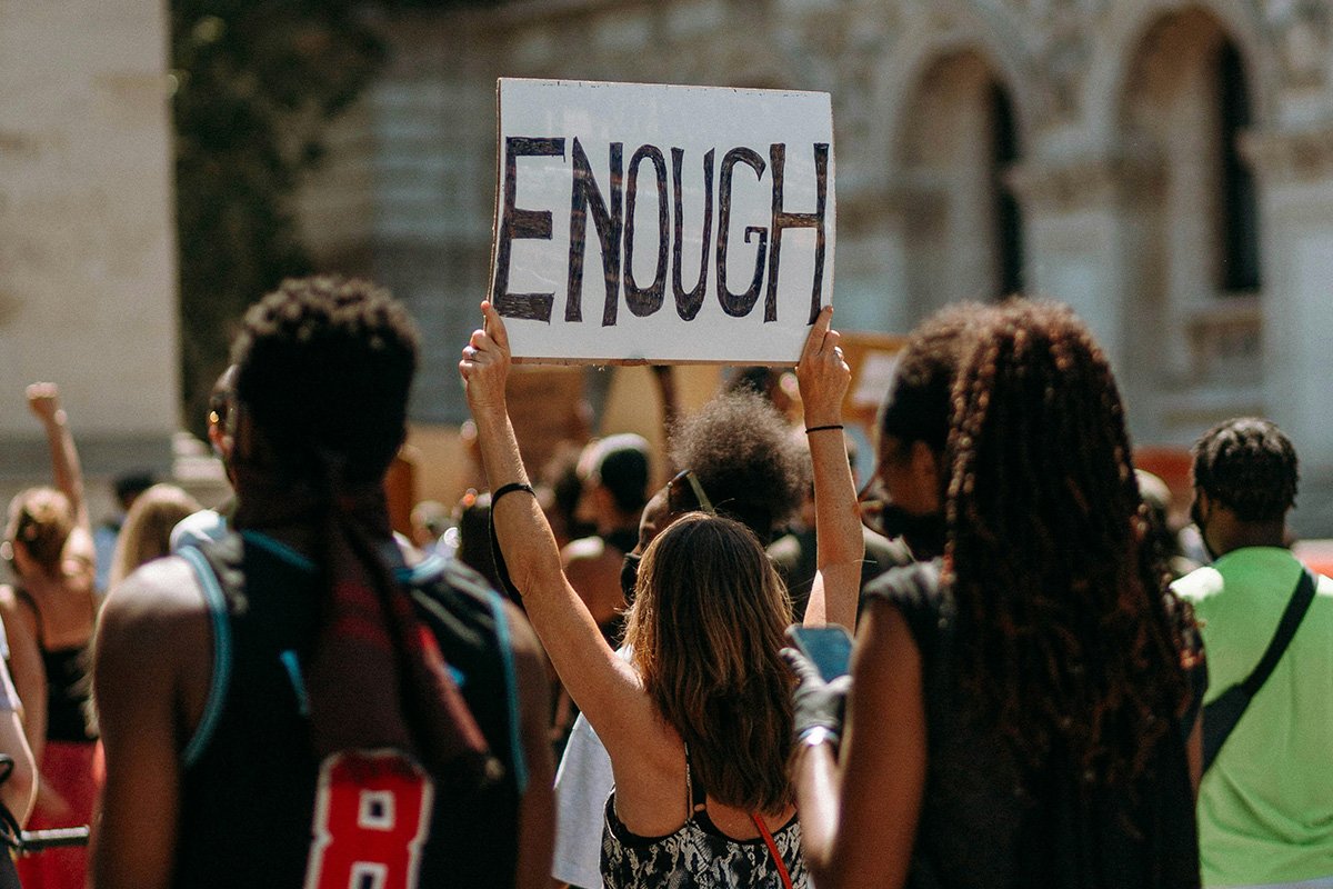 A person holds up a cardboard protest sign reading, “Enough” as she faces away from the camera marching with a group of people.