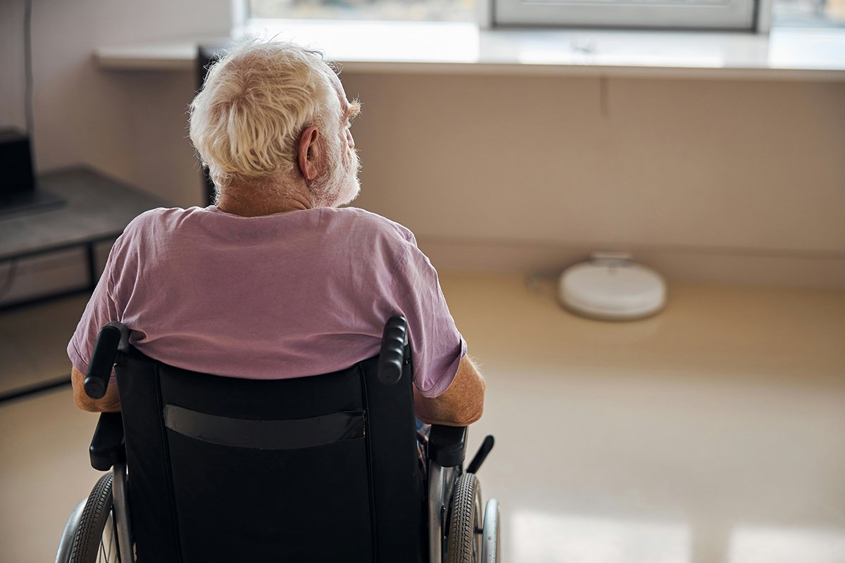 An elderly man with thinning white hair is seated in a wheelchair, facing away from the camera.