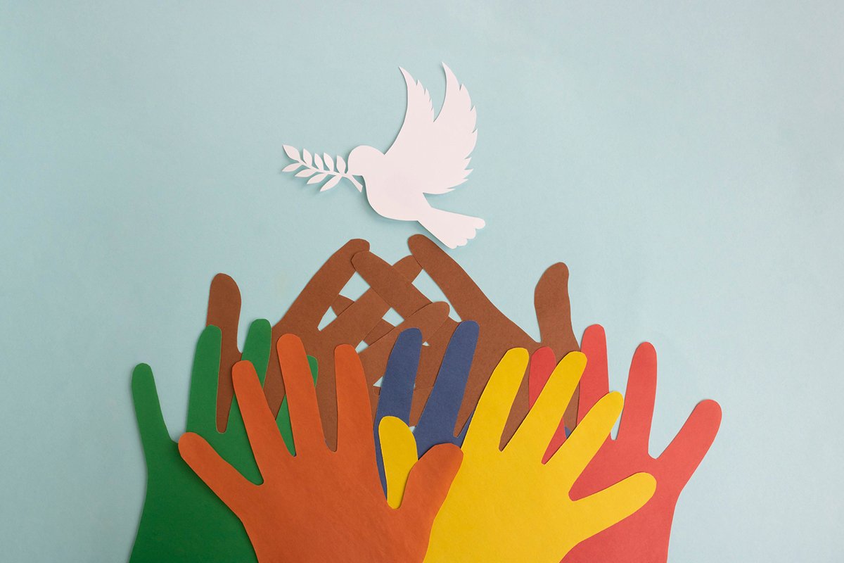 Colored paper hands reach out to a paper dove of peace on a blue background.