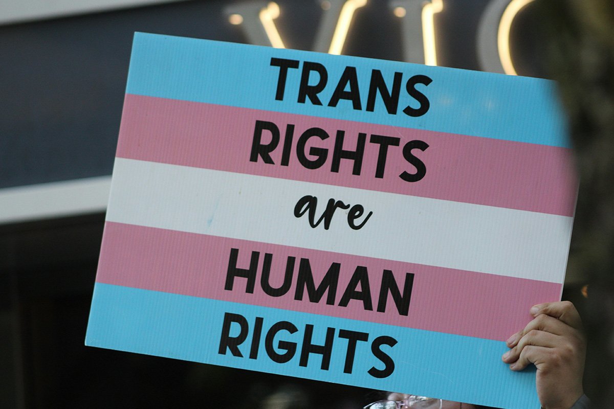 A person holds up a hand-[ainted cardboard sign that reads, “Trans Rights are Human Rights” over a trans flag.