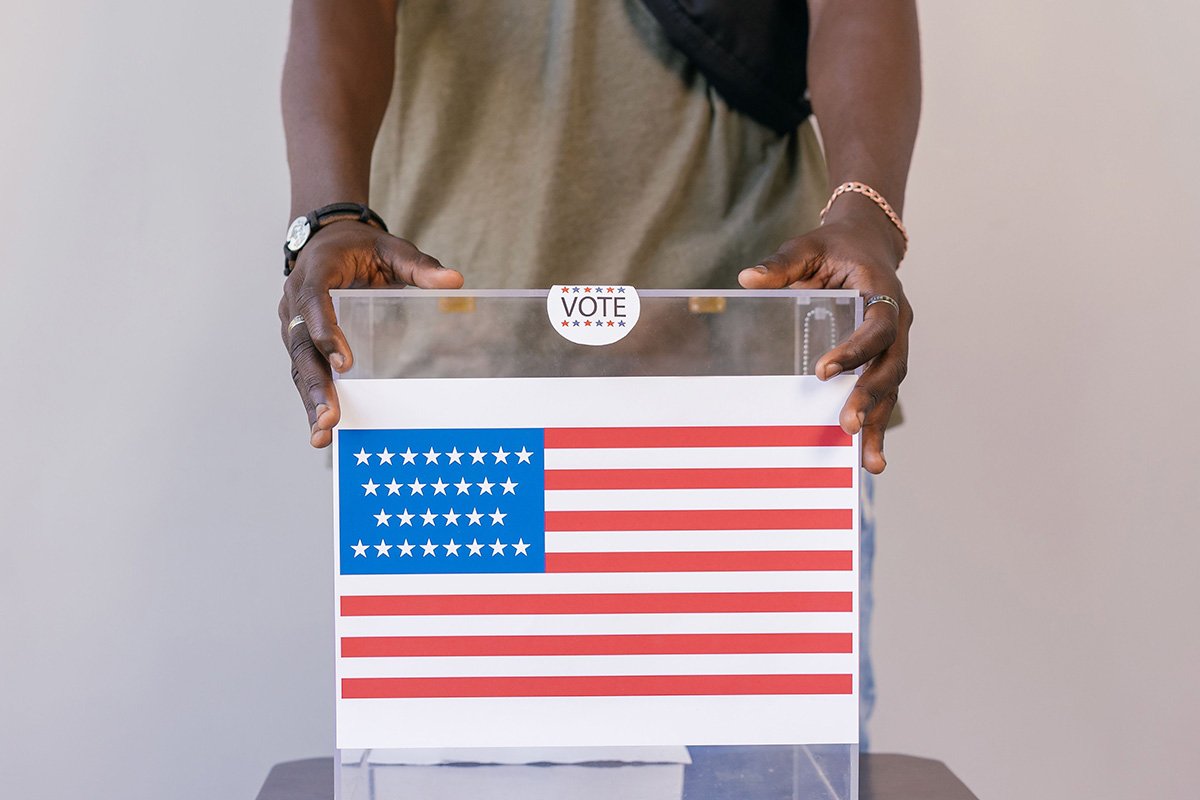 A close-up of a ballot box with an American flag on the front. A Black man’s hands cradle the edges of the box.