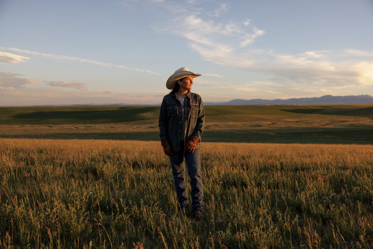 A brunette woman with a demin jacket and a white cowboy hat stands in a grassy field during dusk. The sun casts a warm glow on her relaxed face.
