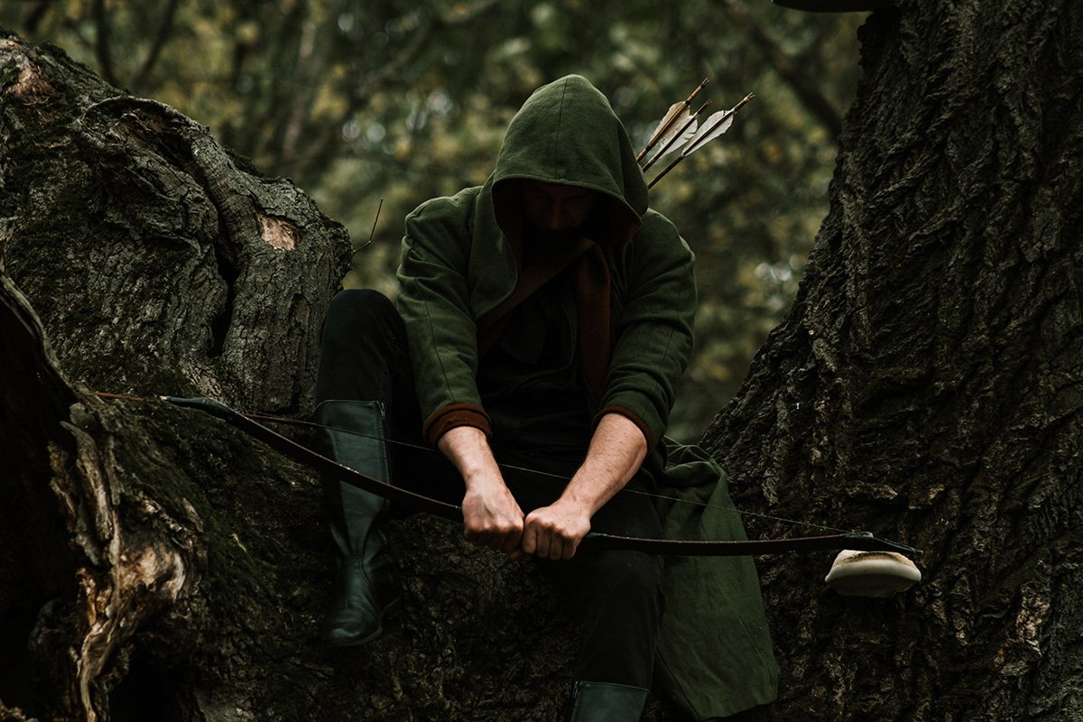 A person sitting in a tree and wearing a Robin Hood costume, where his face is turned down away from the camera, and shielded by his hood. He holds a bow and arrow is his hands.