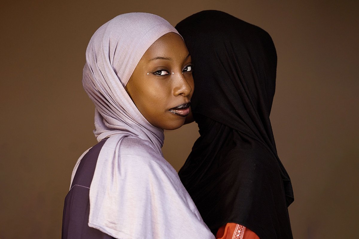 A Black woman in a lavender-colored hijab stands in front of another woman in a black hijab and looks over her shoulder into the camera with a knowing look.