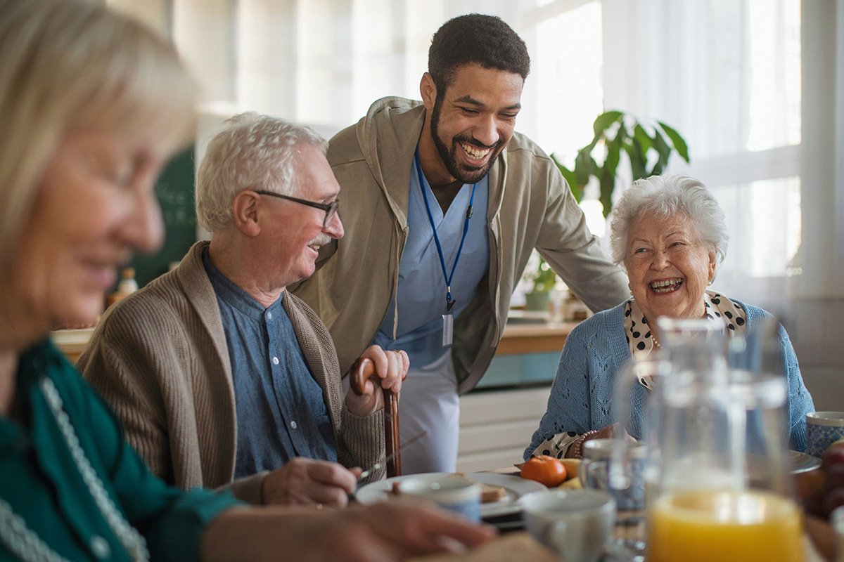 A group of elderly adults sit around a table, smiling and conversing with each other while a Black man in scrubs stands nearby laughing with them.