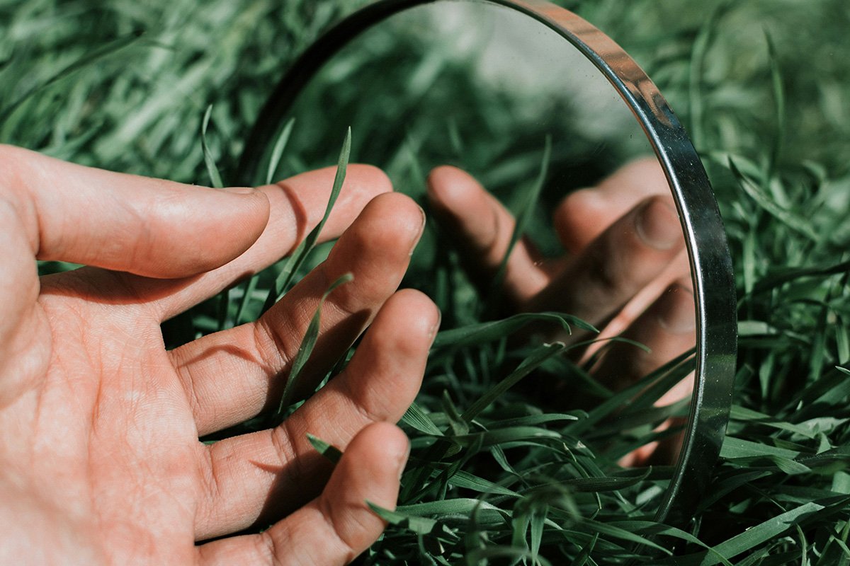 A male hand reaches out to a circular mirror nestled into the grass. The mirror shows a reflection of the hand reaching out.