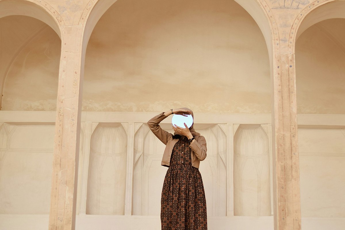 A woman in hijab holds up a mirror to the viewer as she stands between two adobe pillars on an Arab-style building.