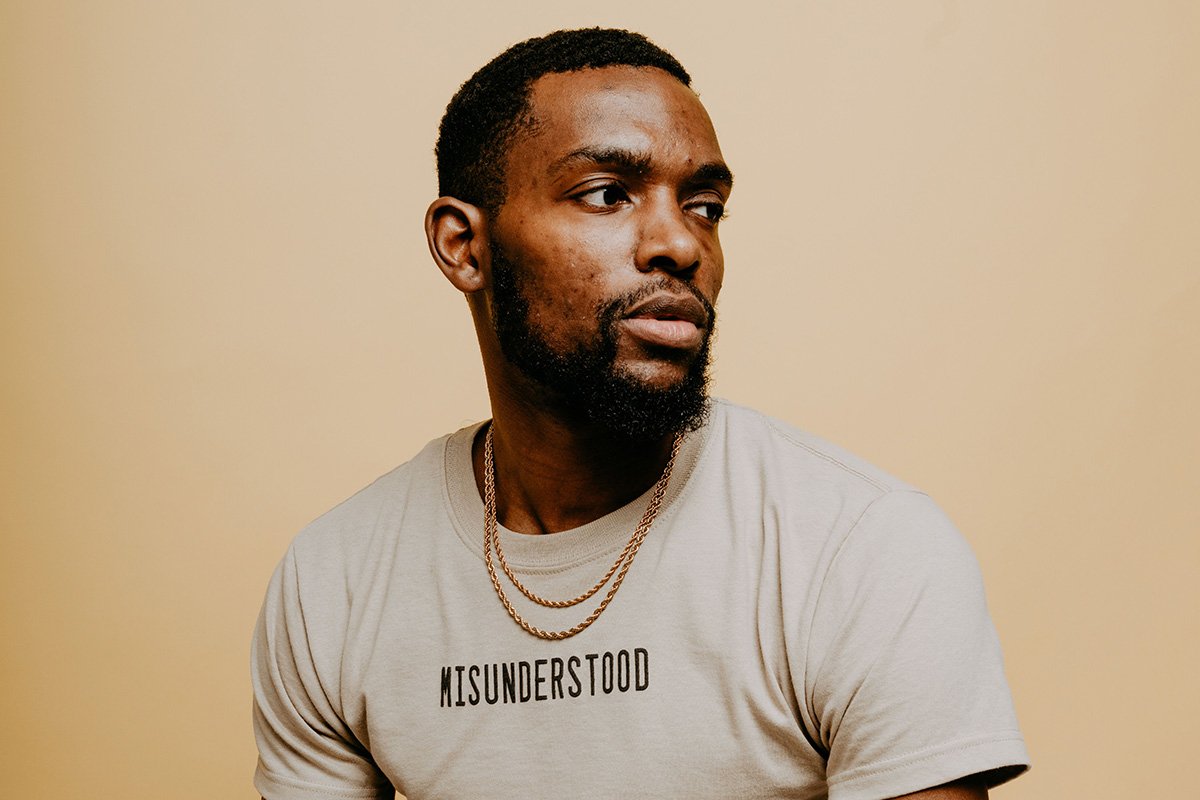 A Black man with cropped hair wears a tan shirt that says “Misunderstood” with two overlapping gold chains. He looks away form the camera.