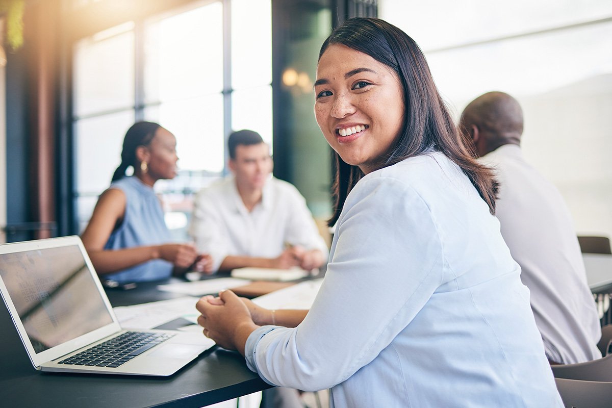 An Asian woman sits in a professional setting at a table with a diverse set of co-workers. She looks over her shoulder with a smile.