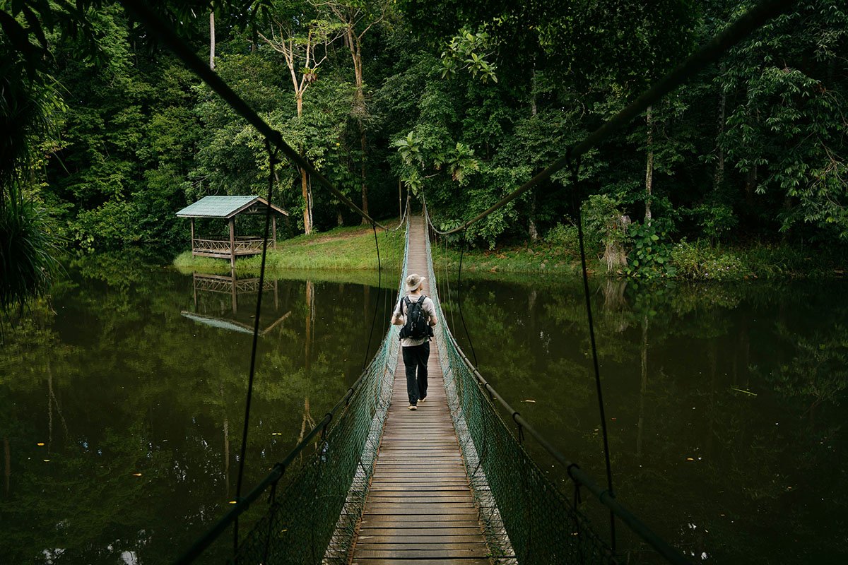 A person in a bucket hat walks over a rope-suspended bridge, hoisted over a body of water.