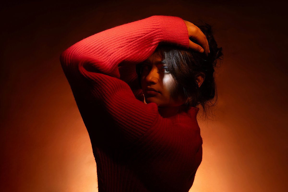 A young South Asian woman with hair pinned up, holding her arm over her head in an oversized red sweater. She looks intently into the camera, with her face in shadow.