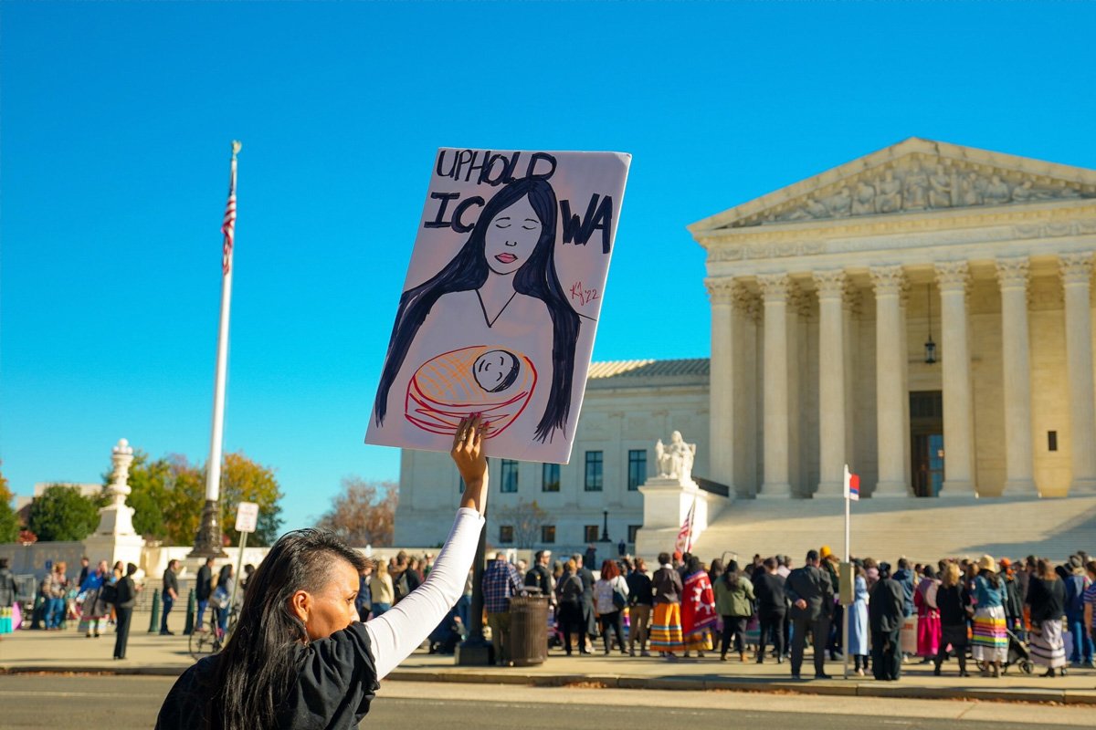 An Indigenous woman holding up a sign that reads “Uphold ICWA” in front of the United State supreme court building in Washington, DC. A symmetrical white marble building with 8 doric columns.