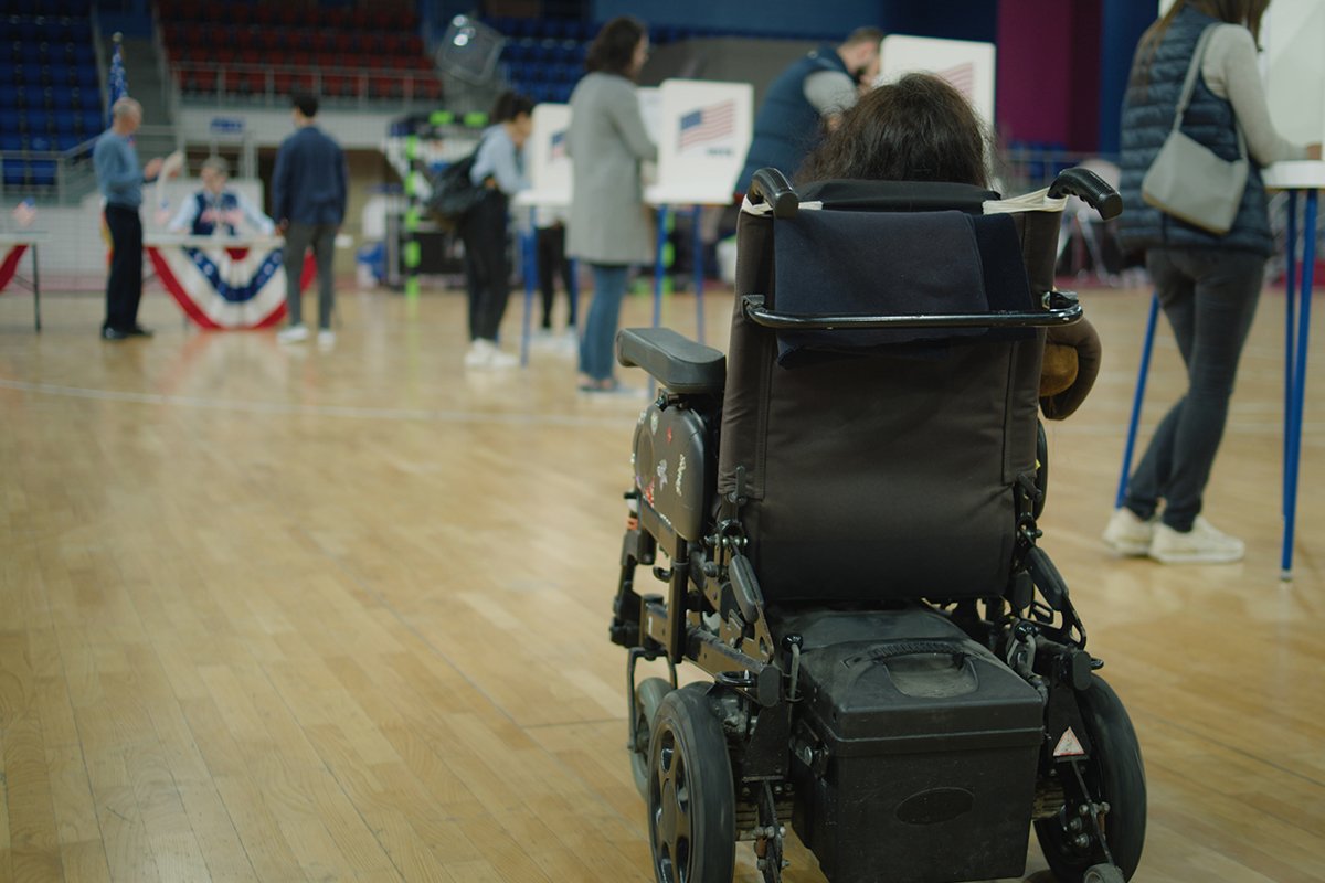 A person in a motorized wheelchair faces away from the camera and towards arranged voting booths in a re-purposed gym.