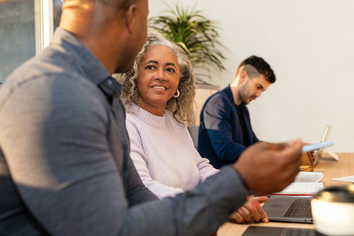 A Black woman with natural gray hair smiles and sits at a table with a computer in front of her, as she talks to a younger coworker.